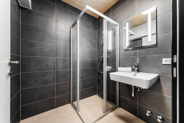 Ein modernes Badezimmer in einem Doppelstudio der International Student Residence der HOK. Ausgestattet mit einer Duschkabine aus Glas. Die Wände sind mit großen, dunkelgrauen Fliesen verkleidet, während der Boden mit hellen Fliesen bedeckt ist. An der rechten Wand befindet sich ein weißes Waschbecken mit einem beleuchteten Spiegel darüber. | © HOK
