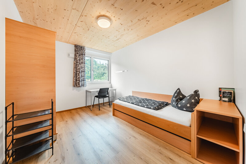 Ein helles, modern eingerichtetes Studierendenzimmer mit einem Einzelbett, einer grauen Bettdecke, einem Schreibtisch mit Stuhl und einem Fenster mit Blick auf grüne Bäume.