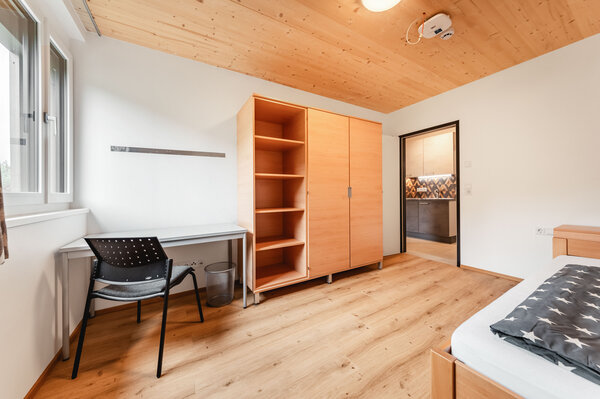 Ein helles, modern eingerichtetes Schlafzimmer mit einem Einzelbett, einer grauen Bettdecke, einem Schreibtisch mit Stuhl und einem Fenster sowie einem großen Schrank. | © HOK