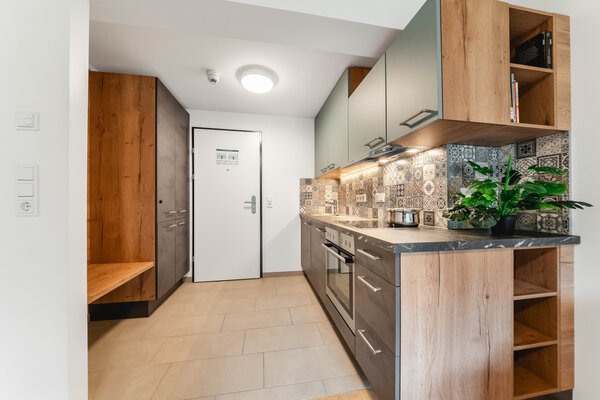 Ein moderner Eingangsbereich mit einer Holzbank. Rechts daneben befindet sich eine moderne Küche mit einer Kombination aus Holz- und dunkelgrauen Schränken. Die Rückwand ist mit gemusterten Fliesen verziert. | © HOK