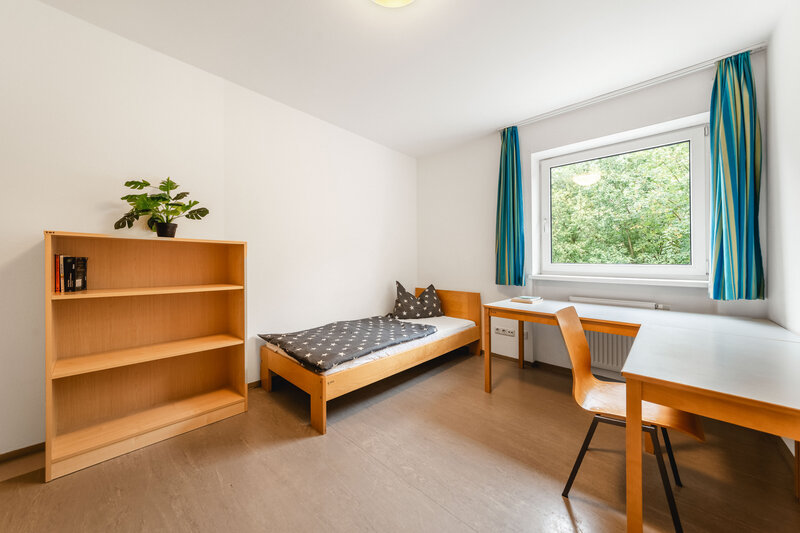Ein hell eingerichtetes Einzelzimmer mit einem Holzbett, einem Schreibtisch und einem Bücherregal.