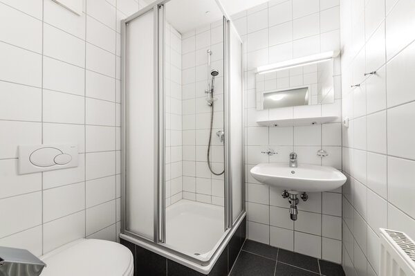 Ein kleines, weiß gefliestes Badezimmer mit einer Duschkabine in der Ecke, einem Waschbecken mit Spiegel und einer Toilette | © HOK