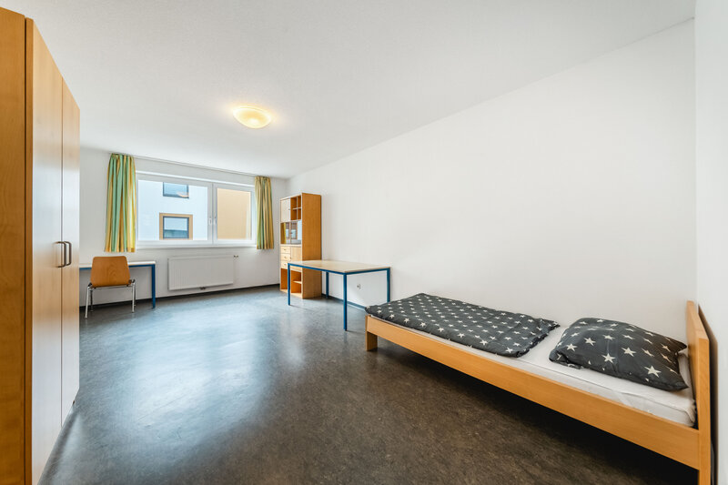 Ein großes Zimmer mit einem Schrank und zwei Tischen. Außerdem gibt es ein großes Fenster und rechts daneben ein Regal.