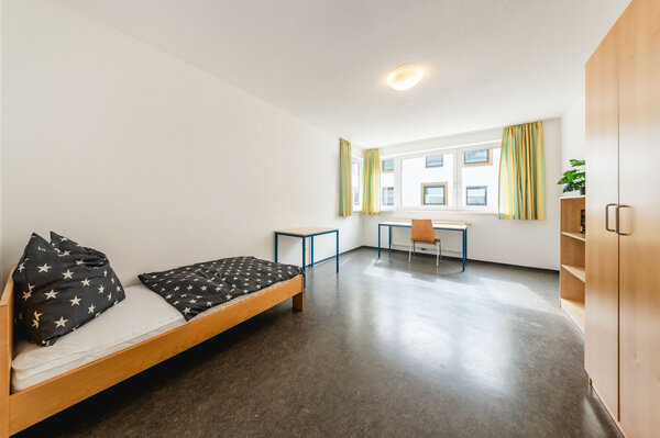 Ein ordentlich eingerichtetes, gut beleuchtetes Zimmer. Links steht ein Einzelbett. Vor dem Bett steht ein kleiner Tisch mit zwei Stühlen. Neben dem Bett rechts befindet sich ein großer Schrank. | © HOK
