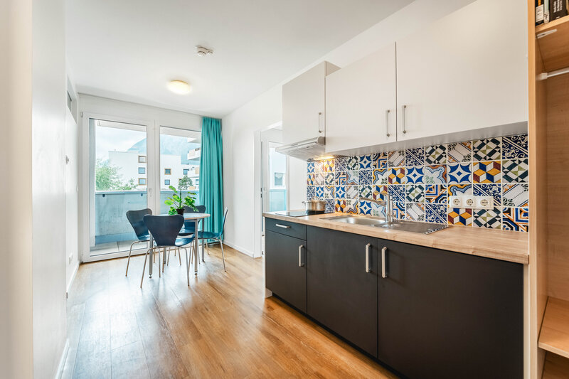 Ein helles, modernes Kücheninterieur mit glatten, dunklen Unterschränken und einer hellen Arbeitsplatte. Im Hintergrund sind ein Tisch, Stühle und ein Balkon zu sehen.