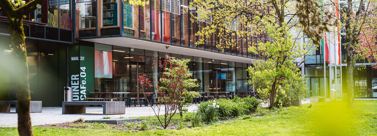 Außenansicht der HOK mit Glasfassade, Cafeteria-Bereich und Frühlingsbepflanzung auf dem Campus