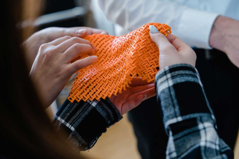 Studierende halten im Makers Lab der HOK ein flexibles, orangefarbenes 3D-gedrucktes Gewebemuster in den Händen.