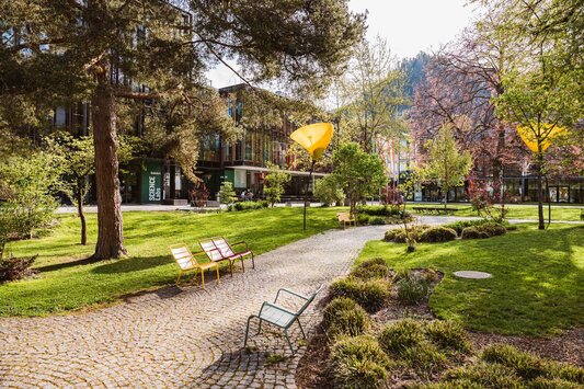 Grüner Campusbereich mit gepflastertem Weg, bunten Bänken und moderner Hochschularchitektur im Hintergrund