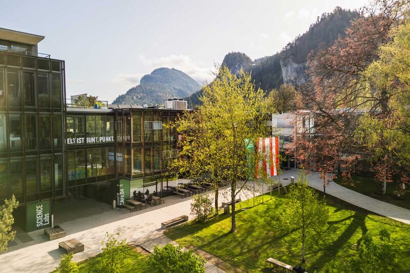 Frühlingshafte Außenaufnahme des Campus der HOK mit Glasfassade, Fahnen und Bergkulisse im Hintergrund