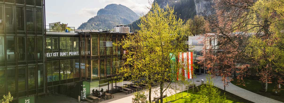 Frühlingshafte Außenaufnahme des Campus der HOK mit Glasfassade, Fahnen und Bergkulisse im Hintergrund