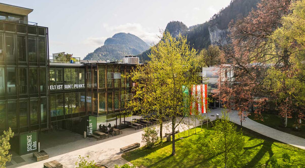 Frühlingshafte Außenaufnahme des Campus der HOK mit Glasfassade, Fahnen und Bergkulisse im Hintergrund
