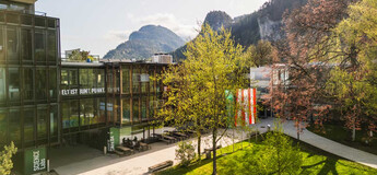 Frühlingshafte Außenaufnahme des Campus der HOK mit Glasfassade, Fahnen und Bergkulisse im Hintergrund