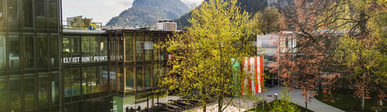 Frühlingshafte Außenaufnahme des Campus der HOK mit Glasfassade, Fahnen und Bergkulisse im Hintergrund
