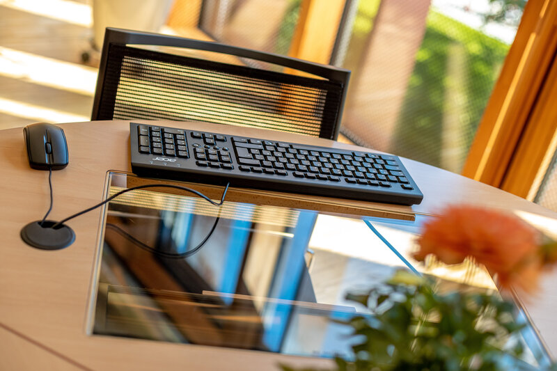 Arbeitsplatz im Lesesaal der kubi mit Tastatur, Maus und Bildschirm auf hellem Holztisch, im Hintergrund Fenster mit Blick ins Grüne.