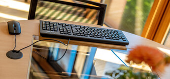 Arbeitsplatz im Lesesaal der kubi mit Tastatur, Maus und Bildschirm auf hellem Holztisch, im Hintergrund Fenster mit Blick ins Grüne.
