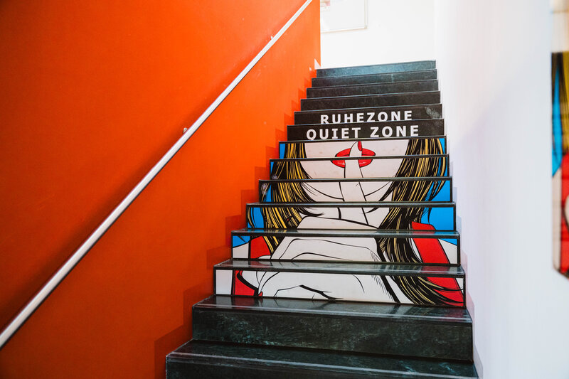 Treppenaufgang zur Ruhezone der kubi mit schwarz-grauen Stufen, auf denen eine Illustration einer Frau mit langem Haar und erhobenem Zeigefinger zu sehen ist, darüber der Text 'RUHEZONE QUIET ZONE', links rote Wand mit weißem Handlauf.