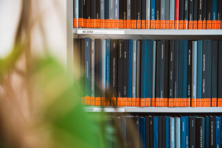 Regal mit gesammelten Masterarbeiten im Lesesaal der kubi, gebundene Bücher mit schwarzen und blauen Einbänden, orangefarbene Etiketten an den Buchrücken.