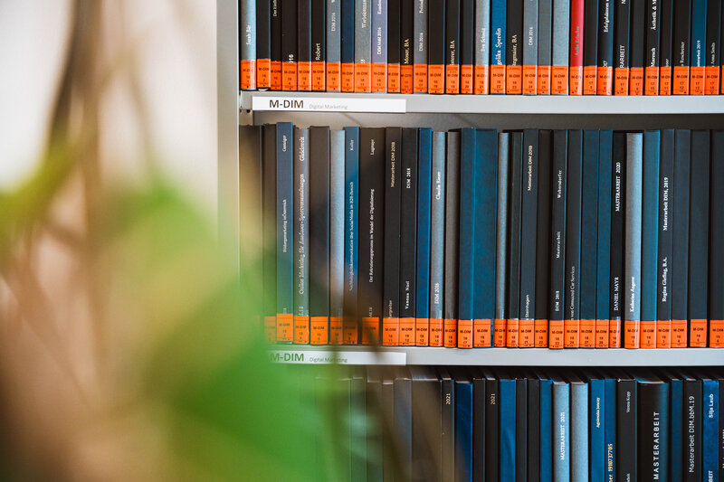 Regal mit gesammelten Masterarbeiten im Lesesaal der kubi, gebundene Bücher mit schwarzen und blauen Einbänden, orangefarbene Etiketten an den Buchrücken.
