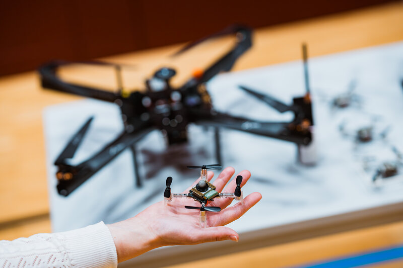 Eine Hand hält eine kleine Quadrocopter-Drohne mit sichtbaren elektronischen Komponenten. Im Hintergrund liegt auf einem Tisch eine größere Drohne.