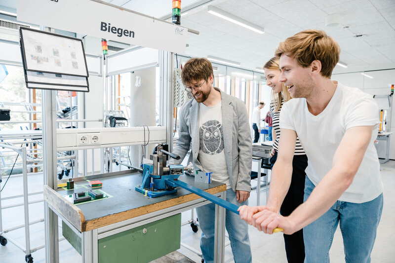 Ein Student und eine Studentin arbeiten gemeinsam mit einem Dozenten an der Biegen-Station im Lean Lab der HOK und wenden Prinzipien des Lean Managements praxisnah an.