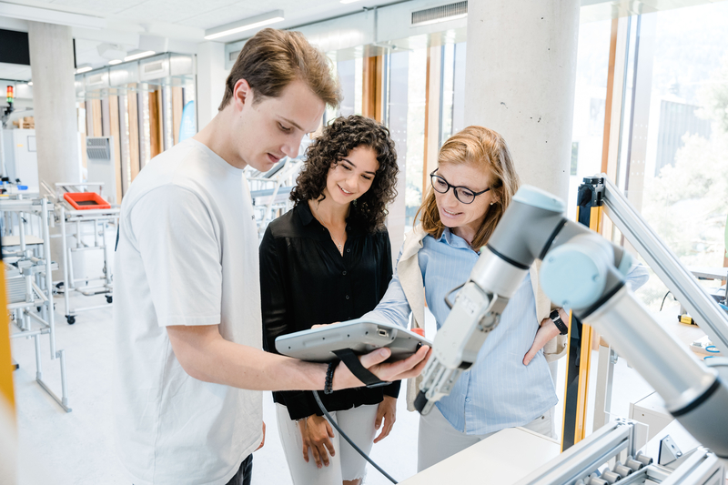 Eine Dozentin der HOK erklärt einer Studentin und einem Studenten im Lean Lab die Steuerung eines kollaborativen Roboters und dessen Einsatz in der Produktionsoptimierung.