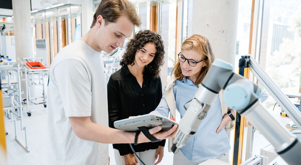 Eine Dozentin der HOK erklärt einer Studentin und einem Studenten im Lean Lab die Steuerung eines kollaborativen Roboters und dessen Einsatz in der Produktionsoptimierung.