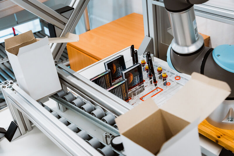 Förderbandanlage mit Robotergreifarm im Lean Lab der HOK, auf dem Materialien automatisiert transportiert und sortiert werden.