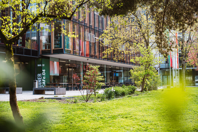 Außenansicht der HOK mit Glasfassade, Cafeteria-Bereich und Frühlingsbepflanzung auf dem Campus