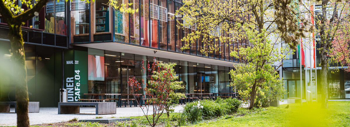 Außenansicht der HOK mit Glasfassade, Cafeteria-Bereich und Frühlingsbepflanzung auf dem Campus