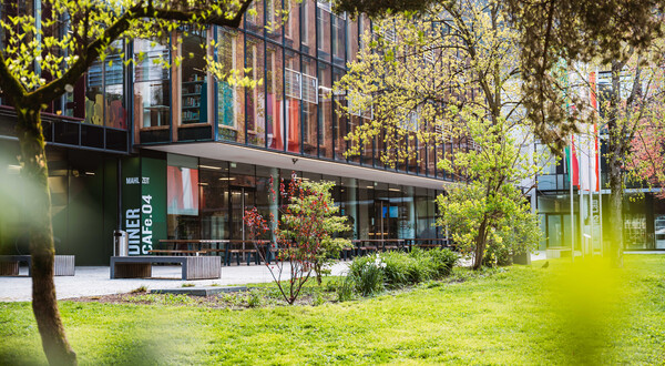Außenansicht der HOK mit Glasfassade, Cafeteria-Bereich und Frühlingsbepflanzung auf dem Campus