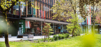 Außenansicht der HOK mit Glasfassade, Cafeteria-Bereich und Frühlingsbepflanzung auf dem Campus