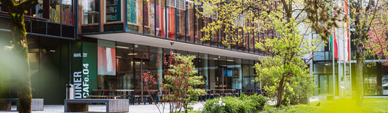 Außenansicht der HOK mit Glasfassade, Cafeteria-Bereich und Frühlingsbepflanzung auf dem Campus
