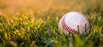 Baseball liegt auf einer grünen Wiese im warmen Abendlicht, mit Fokus auf den Nähten und dem Gras im Vordergrund.