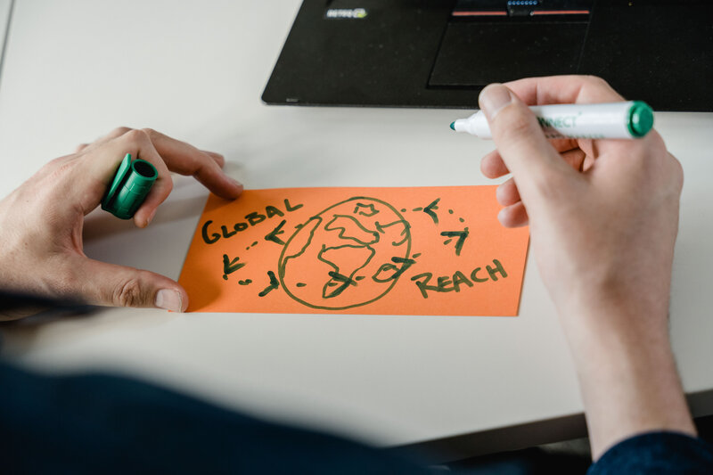 Person zeichnet eine stilisierte Weltkarte mit dem Schriftzug „Global Reach“ – Sinnbild für internationale Vernetzung, Zusammenarbeit und globale Perspektiven.