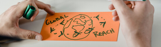 Person zeichnet eine stilisierte Weltkarte mit dem Schriftzug „Global Reach“ – Sinnbild für internationale Vernetzung, Zusammenarbeit und globale Perspektiven.