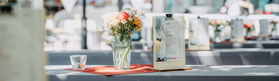 Festlich gedeckter Tisch mit Blumenstrauß, Getränkekarte und Glas unter freiem Himmel – sommerliche Atmosphäre bei einer Veranstaltung.