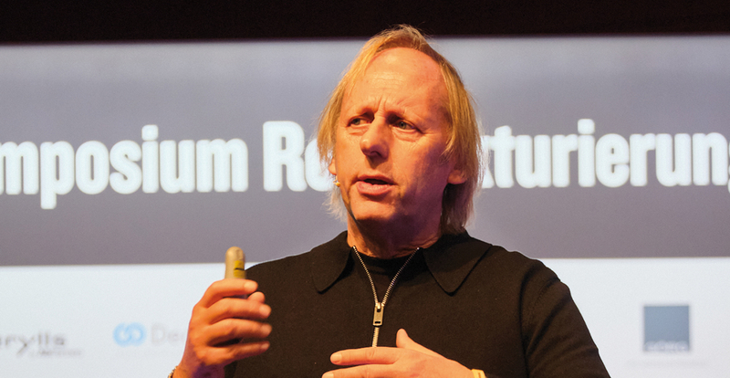 Gastgeber Markus Exler spricht auf der Bühne und hält ein Präsentationsgerät in der Hand, im Hintergrund sind der Schriftzug „Symposium Restrukturierung“ und einige Sponsorenlogos zu sehen.