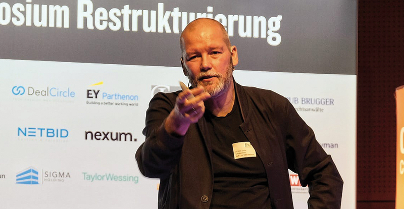 Dr. Stefan Carsten als Speaker auf der Bühne, im Hintergrund Sponsorenlogos und der Schriftzug „Symposium Restrukturierung“