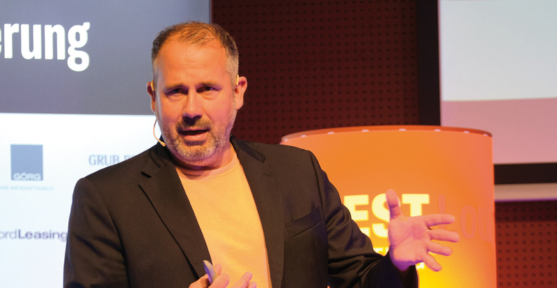 Ulrich Thiem steht auf einer Bühne als Speaker und hält einen Vortrag beim 13. Internationalen Symposium Restrukturierung an der HOK, im Hintergrund sieht man eine Lampe und eine Tafel mit Logos