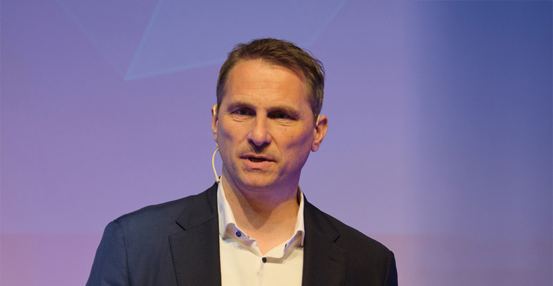 Vortragender Oliver Danninger spricht auf einer Bühne und trägt ein Mikrofon am Ohr, vor einem dezenten, lilafarbenen Hintergrund