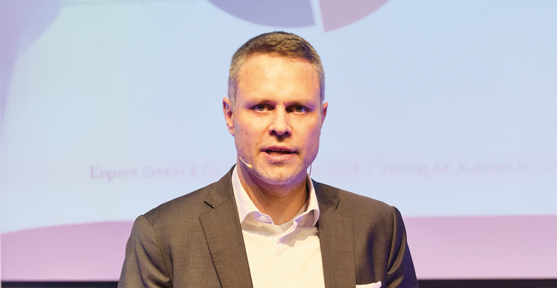 Alexander Kujumdshiev spricht auf der Bühne des 13. Symposiums Restrukturierung, vor einem dezenten, lilafarbenen Hintergrund