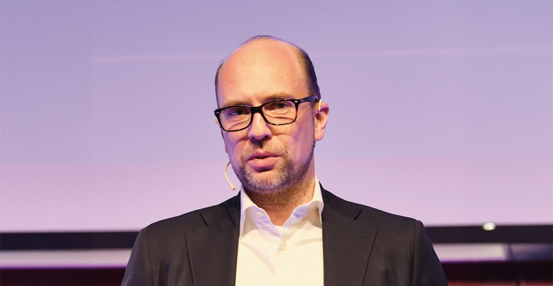 Speaker Philipp Stuetz spricht auf der Bühne beim 13. Internationalem Symposium Restrukturierung der HOK, vor einem dezenten, lilafarbenen Hintergrund