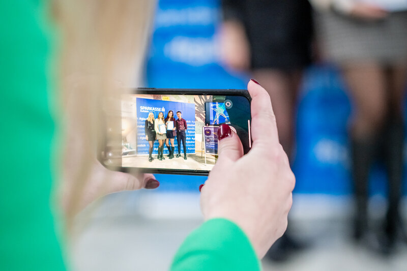 Frauenhände, die ein Mobiltelefon halten, auf dem die Preisträgerinnen des 3. Banking Awards sichtbar sind