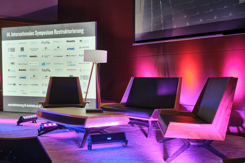 Bühne der 14. Jahreskonferenz Restrukturierung an der HOK, eine moderne Sitzgruppe und mit Tisch und Stehlampe, dahinter eine Aufsteller mit dem Schriftzug 14. Internationales Symposium Restrukturierung und vielen Logos. Die Bühne wird von lila- rosa-farbigen Scheinwerfern angestrahlt
