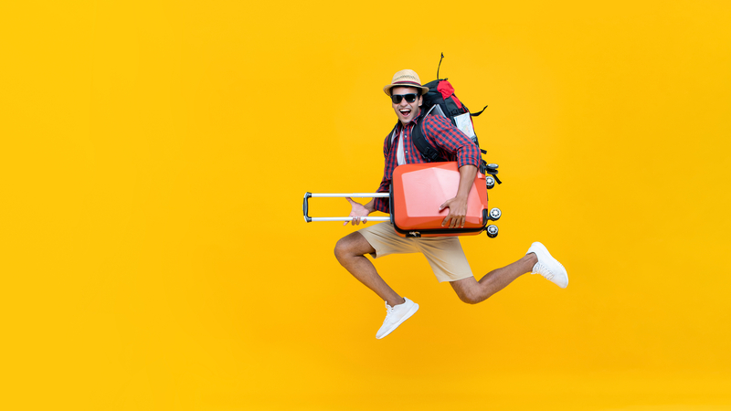 Junger Reisender mit Sonnenbrille, Hut, Rucksack und orangefarbenem Koffer springt vor leuchtend gelbem Hintergrund.