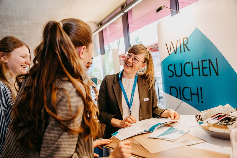 Drei Frauen im Gespräch an einem Messestand mit der Aufschrift "Wir suchen Dich!" im Hintergrund.