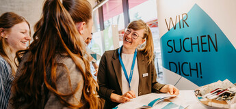 Drei Frauen im Gespräch an einem Messestand mit der Aufschrift "Wir suchen Dich!" im Hintergrund.