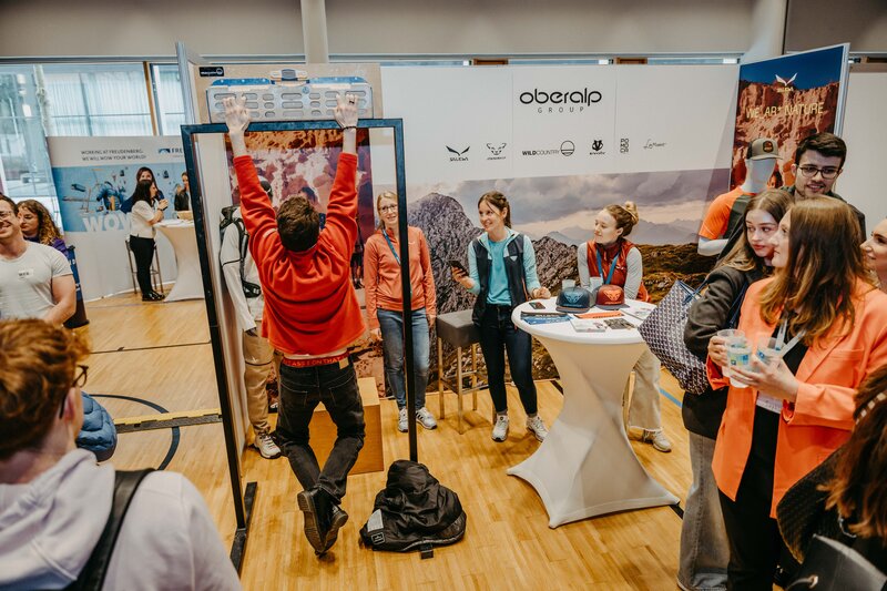 Ein Mann macht Klimmzüge an einem Klettergriffbrett vor dem Oberalp Messestand, während andere Besucher zuschauen und interagieren.