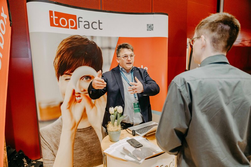 Auf dem Bild ist ein Mitarbeiter der Firma Topfact zu sehen, der einem interessierten Zuhörer etwas erklärt. Der Zuhörer folgt aufmerksam den Ausführungen des Mitarbeiters.