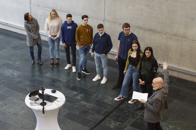 9 Personen stehen in der Aula und halten einen Vortrag.